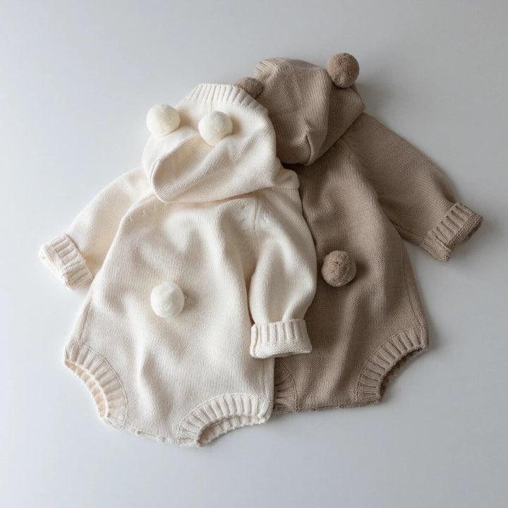 A beige cotton knitted hoodie romper with a teddy bear design, featuring a hood, sleeves, and a front pocket.