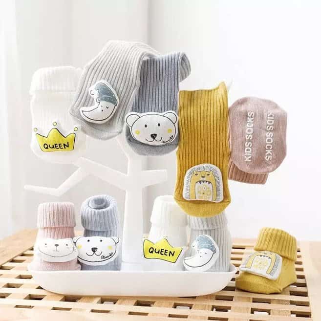 A collection of unisex long cotton socks in various colors, each featuring a cute 3D print design, such as a bear, and adorned with names like 'Queen' and 'King'. The socks are displayed on a tree-like rack with the brand 'Sunshine Baby' visible.
