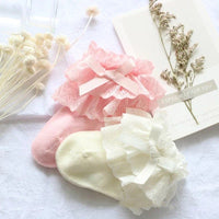 A pair of pink and off-white baby girl lace socks displayed with decorative elements like a card and a sprig of dried flowers in the background.