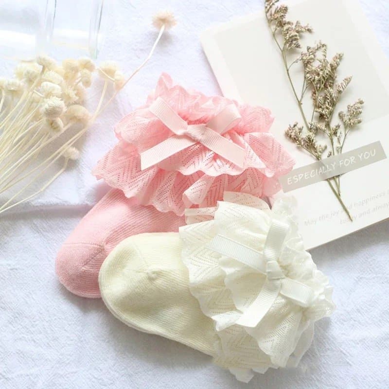 A pair of pink and off-white baby girl lace socks displayed with decorative elements like a card and a sprig of dried flowers in the background.